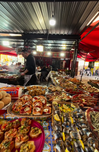 A vendor tending a Palermo street-food market stall spread with bruschette, sardines and fried snacks